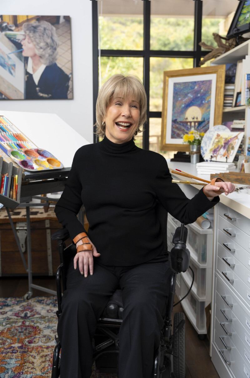 Joni Eareckson Tada smiling in her wheelchair in her art studio, holding paintbrushes.