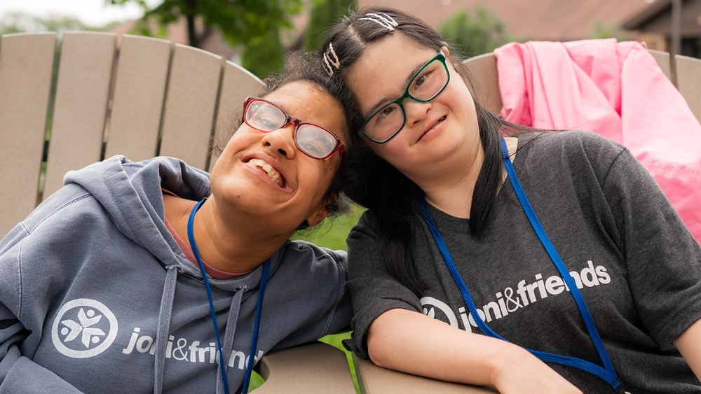 Two young women in Joni and Friends shirts and lanyards resting their heads together and smiling.